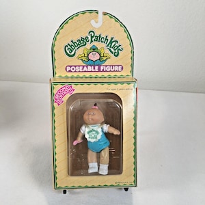 May include: A Cabbage Patch Kids poseable figure in its original packaging. The doll has a round face, a small tuft of dark hair, and wears a white bib with green print over a blue outfit and white shoes. The box features the text Second Edition.