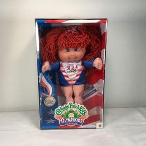 May include: A Cabbage Patch Kids Olympikids special edition doll with red yarn hair, blue eyes, and a light skin tone. The doll wears a blue, white, and red leotard featuring the USA text and Olympic rings logo, packaged in a box with flag graphics.