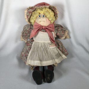 Pode incluir: Uma boneca de trapo vintage com um rosto macio e redondo, olhos azuis e cabelo encaracolado amarelo. A boneca usa um vestido com estampa floral, um avental branco e um laço rosa. Os sapatos da boneca são pretos e ela usa um chapéu.