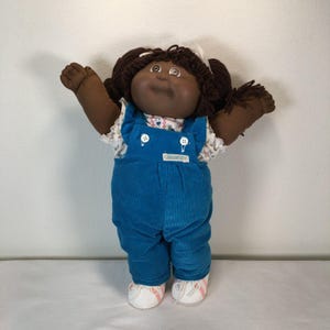 Może przedstawiać: Lalka Cabbage Patch Kids z ciemnobrązowymi włosami z włóczki w kucykach, ciemną karnacją i brązowymi oczami. Lalka ma na sobie niebieskie sztruksowe ogrodniczki z białymi guzikami, białą koszulę w kwiaty oraz białe buty z różowymi paskami.