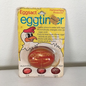 May include: A red egg-shaped kitchen timer in its original yellow cardboard packaging. The packaging features a cartoon chicken illustration and text including Eggsact eggtimer, Hammerhead Products, and instructions for soft, medium, and hard eggs.