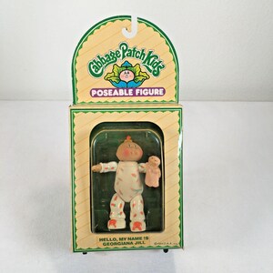 Puede incluir: Una figura articulada de Cabbage Patch Kids en su empaque original. La muñeca, llamada Georgiana Jill, viste un mameluco blanco con estampados de zanahorias naranjas y sostiene un pequeño juguete color canela. La caja tiene un borde verde.