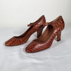 May include: A pair of brown, textured leather pumps with a square toe and a strap across the top of the foot. The shoes have a medium-height heel and a classic, vintage design. The leather has a reptile-like pattern.