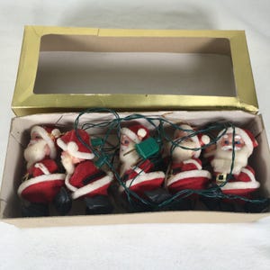 Puede incluir: Una cadena de cinco luces navideñas de Papá Noel en una caja de cartón. Cada figura de Papá Noel tiene un traje rojo, barba blanca y botas negras. Las luces están ensartadas en un cable verde.