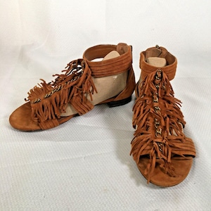 May include: A pair of brown suede sandals with fringe and gold chain accents. The sandals have a flat sole and a back zipper. The fringe detail is prominent across the top of the foot and around the ankle.
