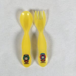 May include: A yellow plastic spoon and fork set. The spoon and fork have rounded handles and feature a colourful graphic of a cartoon train. The spoon has the name "James" on it.