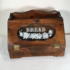 Könnte beinhalten: Ein dunkelbrauner Brotkasten aus Holz mit einer aufklappbaren Fronttür. Die Tür verfügt über ein zentrales Glaselement, das mit weißen Rosenmotiven und dem Wort BREAD in weißer Schrift verziert ist, befestigt mit zwei messingfarbenen Scharnieren.