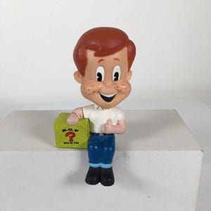 May include: A cartoon-style figurine of a boy with reddish-brown hair and freckles, wearing a white shirt and blue pants. The figure is sitting and holding a lime green cube with the text B.O.F. and BOX OF FUN printed above a red question mark.