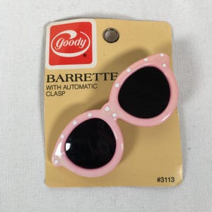May include: A pink barrette in the shape of sunglasses with black lenses and white polka dots. The barrette is attached to a tan cardboard backing with the brand name "Goody" and the text "Barrette with Automatic Clasp."