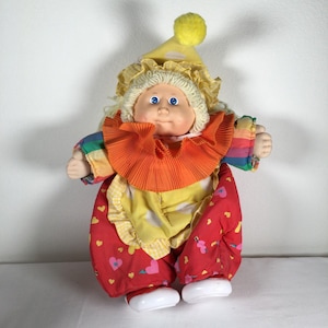 May include: A vintage clown doll with a yellow hat and a ruffled orange collar. The doll has a painted face with blue eyes and is wearing a colorful outfit with red pants decorated with hearts. The doll's hair is blonde.