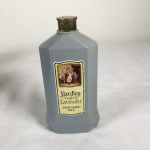 May include: A vintage Yardley English Lavender perfumed talc bottle. The gray, geometric bottle has a gold-colored cap and a label with an illustration and text. The label reads "Yardley English Lavender Perfumed Talc."