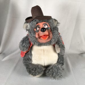 Könnte beinhalten: Ein Vintage-Plüschtier eines bärenartigen Charakters mit braunem Hut und roter Weste. Das Spielzeug hat einen grauen Fellkörper, ein pfirsichfarbenes Gesicht und eine schwarze Nase. Der Mund des Bären ist geöffnet und zeigt eine orangefarbene Zunge.