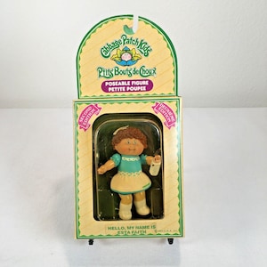 May include: A Cabbage Patch Kids poseable figure in original packaging. The doll has brown hair, a teal dress with a white apron, and white shoes. The packaging features the text Cabbage Patch Kids, Second Edition, and Hello, My Name Is Esta Faith.