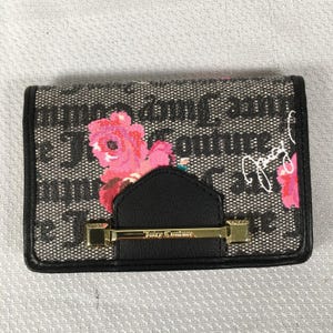 May include: A black and white wallet with a floral design. The wallet features a black flap with a gold-coloured clasp and the words "Juicy Couture" in gold. The wallet has a black border.