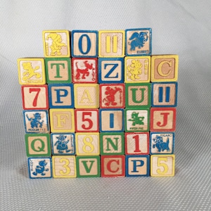 May include: A stack of colorful wooden alphabet blocks. Each block features a letter, number, or cartoon character illustration. The blocks are painted with primary colors and have yellow, blue, red, and green accents. The blocks are arranged in a square formation.