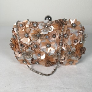 Pode incluir: Uma clutch floral com uma alça de corrente prateada. A bolsa retangular é coberta por flores de tecido pêssego e branco, com detalhes de cristal prateado e transparente. Fecho de bola prateada.