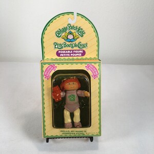 Puede incluir: Una figura articulada de Cabbage Patch Kids en su embalaje original. La muñeca tiene el pelo rojo, una camiseta lavanda y pantalones amarillos, y sostiene un pequeño oso marrón. La caja incluye el texto Cabbage Patch Kids, P'tits Bouts de Choux y Jennifer Ethel.