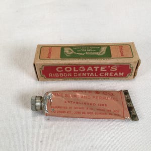 Könnte beinhalten: Eine Vintage-Pappschachtel von Colgate's Ribbon Dental Cream und eine passende Metalltube. Die Schachtel zeigt grünen und roten Text mit dem Satz 'Comes out a ribbon lies flat on the brush'. Die Tube ist rosa mit rotem Text über Colgate & Co.