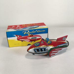 Peut inclure: Un jouet en fer blanc en forme de fusée de course rouge, blanche et verte avec un pilote au casque bleu, portant l'inscription "ROCKET RACER" et le chiffre "6". Il est accompagné d'une boîte en carton rectangulaire aux graphismes assortis.