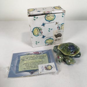May include: A ceramic turtle figurine with a green and blue shell, accompanied by its original cardboard box featuring floral and butterfly motifs, and a registration of ownership card in a clear plastic sleeve with the text Clayworks by Heather Goldminc.