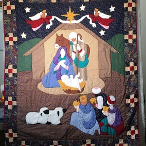 May include: A rectangular fabric quilt featuring a nativity scene with a central stable, a star, and angels. The scene includes figures of a person in blue, a person in striped robes, a baby in a manger, sheep, and three figures in colourful robes.