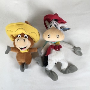 Puede incluir: Dos peluches que representan un burro marrón con sombrero amarillo y pañuelo amarillo, y un caballo blanco con sombrero de vaquero rojo, cinturón rojo con hebilla, pañuelo azul claro y una insignia de estrella amarilla en el pecho.