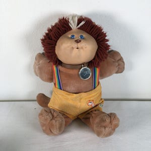 May include: A vintage plush toy featuring a lion-like design with brown fur and a cream-colored face. It has blue eyes, a brown collar with a silver tag, and rainbow suspenders over yellow corduroy shorts. The toy is approximately 10 inches tall.