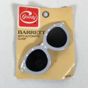May include: A light gray barrette in the shape of sunglasses with black lenses and white polka dots. The barrette is on a yellow card with the Goody logo and the text "BARRETT WITH AUTOMATIC CLASP". The card has a hole at the top.