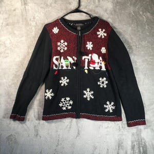 May include: Black zip-up sweater with a festive Christmas design. The sweater features the word "SANTA" with a Santa Claus graphic, snowflakes, and colorful lights. The top portion is red with white snowflake accents.
