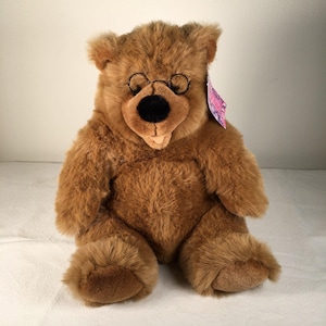 May include: A brown plush teddy bear with a black nose and small round glasses. The bear has a tag attached to its ear. The bear is sitting with its legs extended.