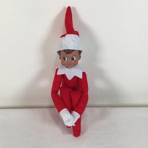 Peut inclure: Une poupée elfe articulée avec une peau foncée, des yeux bleus et des joues roses. La poupée porte un costume rouge vif avec un col blanc et un chapeau rouge assorti doté d'un bord blanc. Les mains et les pieds sont recouverts de tissu blanc.