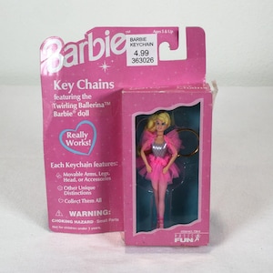 May include: A Barbie Twirling Ballerina keychain in original pink packaging. The doll features blonde hair, a silver bodice, and a pink tulle skirt. The packaging includes text such as Barbie, Key Chains, Really Works, and a warning about small parts.