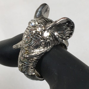 May include: Silver-toned elephant ring with clear rhinestone accents. The elephant's head is detailed with textured lines and features large ears and a trunk. The ring is worn on a black surface.