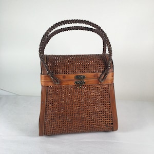 May include: A handcrafted brown woven bamboo handbag with a rectangular shape and a hinged lid. The bag features a woven pattern, a clasp closure, and a braided handle. The bag is designed for carrying small items.