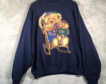 Op de afbeelding: Marineblauwe sweatshirt met een afbeelding van drie teddyberen in een rotan schommelstoel. De beren dragen geruite en gebreide kleding. De sweatshirt heeft lange mouwen en een ronde hals.