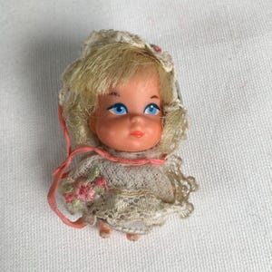 May include: A small vintage doll with blonde hair and blue eyes. The doll wears a white lace dress and a matching bonnet. A pink ribbon is around the neck and a small bouquet of pink flowers is on the dress. The doll is a collectible item.