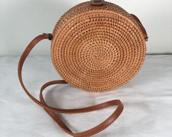 Op de afbeelding: Een ronde crossbodytas van gevlochten rotan met een natuurlijke lichtbruine afwerking. De tas heeft een cirkelvormig, gestructureerd patroon dat vanuit het midden uitstraalt, een bruine leren riem en een kleine leren sluiting aan de bovenkant.