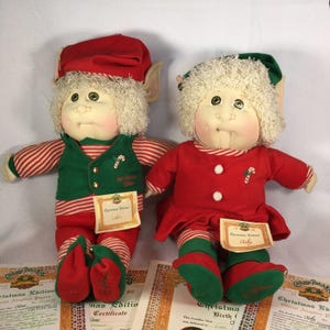May include: Two Cabbage Patch Kids dolls dressed as elves for Christmas. The boy elf is wearing a red and white striped shirt, a green vest, and a red hat with white trim. The girl elf is wearing a red dress with white stripes, a green vest, and a green hat with white trim. Both dolls have white fur and green eyes.