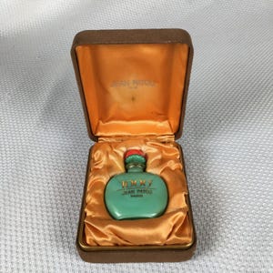 May include: A vintage teal perfume bottle with a red stopper, labelled "1000 JEAN PATOU PARIS," is presented in an open brown velvet-lined box with a gold satin interior. The bottle is the focal point of the image.