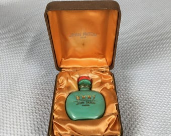 Könnte beinhalten: Eine Vintage-Parfümflasche in Türkis mit rotem Verschluss, beschriftet mit "1000 JEAN PATOU PARIS", wird in einer offenen, braunen, samtgefütterten Schachtel mit goldenem Satin-Innenraum präsentiert. Die Flasche ist das Hauptmerkmal des Bildes.