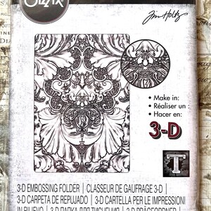 May include: A package for a Sizzix 3-D Texture Fades A6 embossing folder. The package features a detailed, ornate design in black and white. The package also includes the text "3-D TEXTURE FADES" and the name "Jim Holtz".