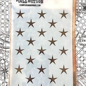 May include: A stencil with a star pattern, featuring dark brown stars on a light blue background. The stencil is labeled "STAMPERS ANONYMOUS" and "Jim Holtz COLLECTION". The stencil is designed for layering and includes the text "STENCIL".