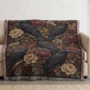 May include: A woven throw blanket featuring a dark pattern of black ravens interspersed with large, detailed flowers in shades of burgundy, cream, and muted gold, finished with a fringed edge along the top and bottom of the rectangular textile.