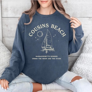 May include: A navy blue sweatshirt with the words "COUSINS BEACH" in a curved arch, with a sailboat graphic. Below the graphic, the text reads "MASSACHUSETTS SUMMER UNDER THE MOON AND THE STARS". The sweatshirt is a relaxed fit.