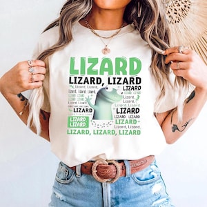 Op de afbeelding: Crèmekleurig T-shirt met het woord "LIZARD" in grote groene letters, tekst over hagedissen en een cartoon hagedis. Het shirt wordt gedragen met een blauwe spijkerbroek en een bruine riem.