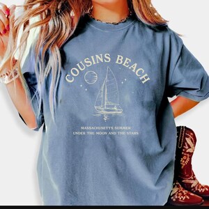 May include: A blue t-shirt with the words "COUSINS BEACH" arched across the top, and a sailboat graphic. Below the graphic, the text reads "MASSACHUSETTS SUMMER UNDER THE MOON AND THE STARS".