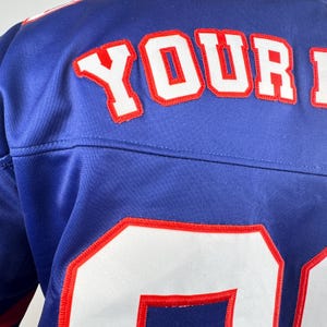 May include: Close-up of a blue jersey with white and red lettering and numbers. The word "YOUR" is visible in large, white letters with red trim. The number "0" is also visible.