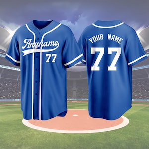 May include: A blue baseball jersey with white accents, featuring the text "Anyname" and the number 77 on the front. The back of the jersey displays "YOUR NAME" and the number 77. The jersey is displayed on a baseball field.