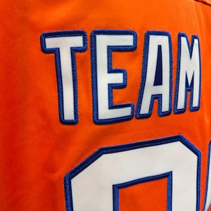 May include: Close-up of an orange jersey with the word "TEAM" and the number "0" stitched in white with blue trim. The jersey has a textured surface and a visible seam.
