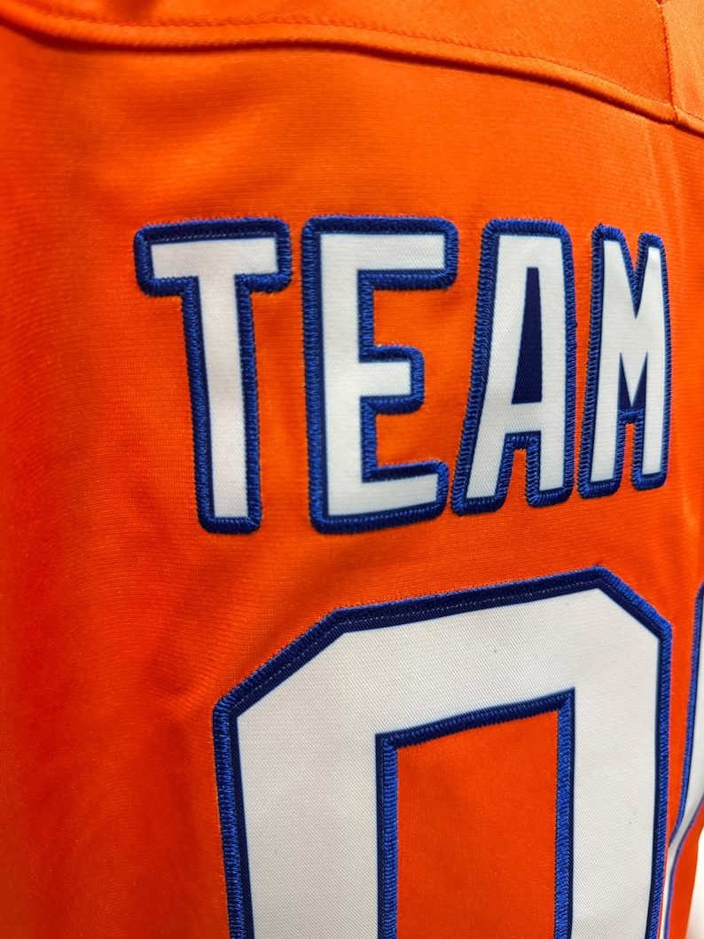 May include: Close-up of an orange jersey with the word "TEAM" and the number "0" stitched in white with blue trim. The jersey has a textured surface and a visible seam.